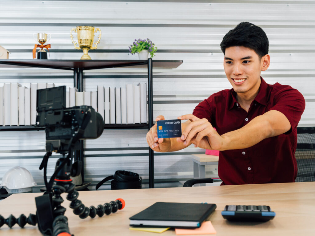 Smiling Asian male recording video selling stuff through social media and video platform showing credit card using camera sitting at table in house