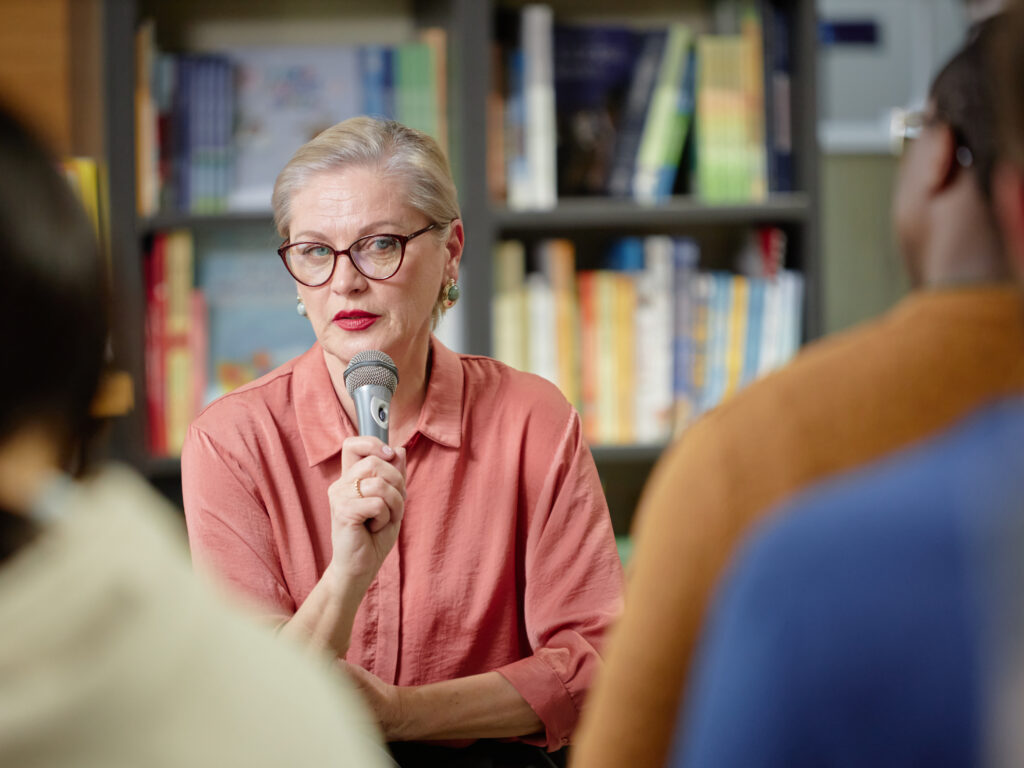 Caucasian senior woman holding microphone speaking to diverse group of people in library setting, wearing glasses and looking attentive, engaging audience during discussion