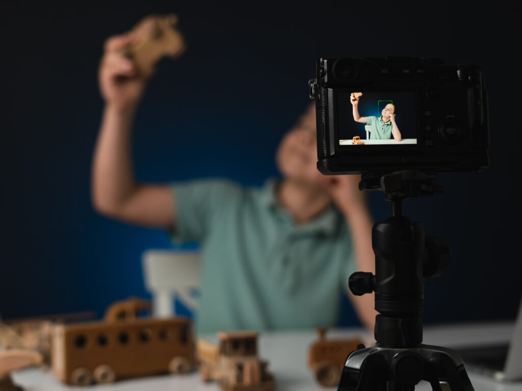 Kid blogger filming video on camera indoors with blue light in background. Online reviewing. Promoting eco wooden toys. Kid recording his gameplay for vlog.